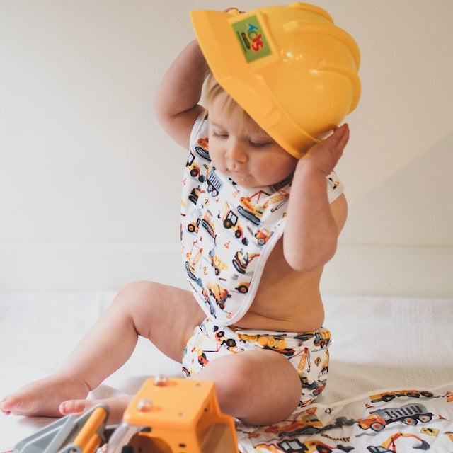 Baby wearing a yellow hard hat sitting on a diggers play mat and wearing a matching bib and nappy