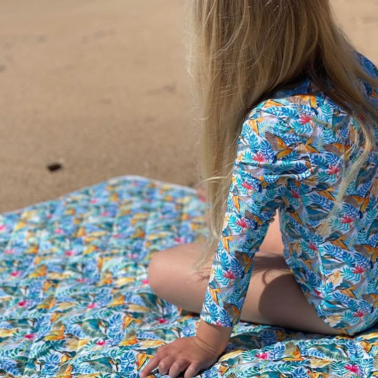 Little girl sitting on a kingfishers play mat on a beach wearing a matching sun safe swimsuit